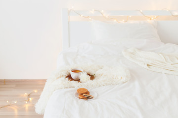 Cup of tea, white flower, wooden tray, cookies, fluffy plaid, knitted sweater and christmas garland lights on white bedding at home. Breakfast in bed. Winter homely scene. Cozy modern white bedroom.