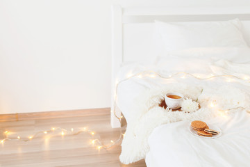Cup of tea, white flower, wooden tray, cookies, fluffy plaid, knitted sweater and christmas garland lights on white bedding at home. Breakfast in bed. Winter homely scene. Cozy modern white bedroom.