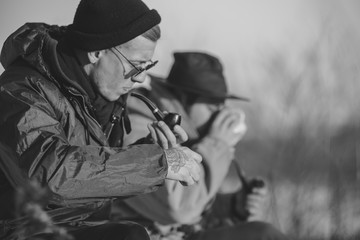 Men is smoking tobacco-pipe and drink coffee. Outdoor portrait of hikker in black sunglasses sitting outdoors during sunny winter day.