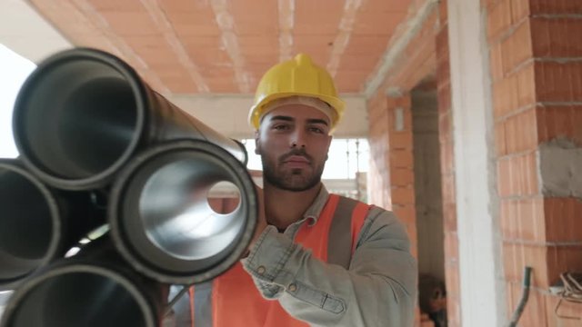 Proud people working in construction site. Portrait of happy white man at work in new house inside apartment building. Professional latino worker carrying pipes and smiling at camera. Slow motion