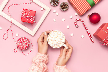 Female hands in knitted sweater holding cup of marshmallows and Christmas candy cane gifts boxes red ball photo frame on pink background top view Flat Lay. Winter traditional drink food. Festive decor