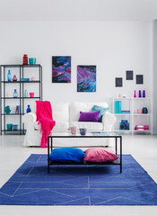 Table with cushions on blue carpet in living room interior with posters above white sofa. Real photo