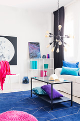 Table on blue carpet in apartment interior with white couch and pink blanket on chair. Real photo