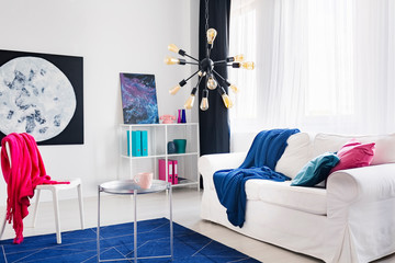 Blue blanket on white sofa and pink cloth on chair in living room interior with posters. Real photo