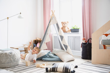 Smiling kid on the floor and plush toy in bedroom interior with pouf and tent next to lamp. Real...