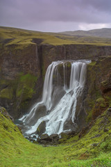 The amazing waterfall Fagrifoss shortly after the rain.  waterfalls in Iceland. Picturesque waterfall in the beautiful valley. In the foreground are stones and rocks with green vegetation. Sky is dark