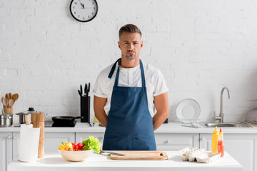 confident man in apron standing on the kitchen and looking at camera