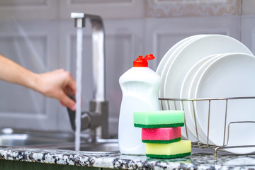 Different clean plates in dish drying rack, dish sponges and dishwashing detergent on the table on kitchen counter. Washing dirty dishes