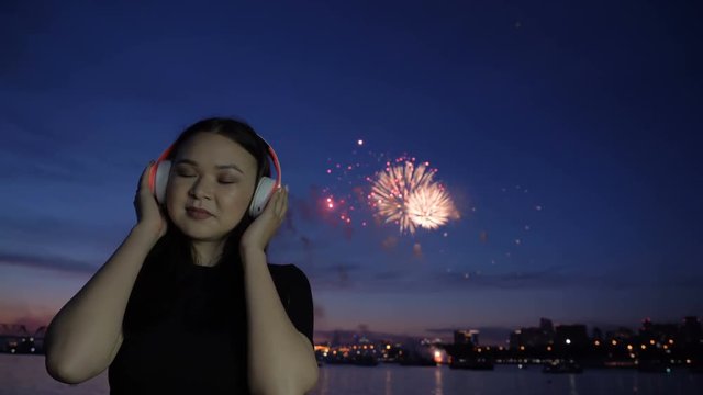 One young woman smile and listening music at headphones at blur scene with exploding fireworks at horizon slow motion. Free 20s female person enjoying alone at night on the holiday on the town beach