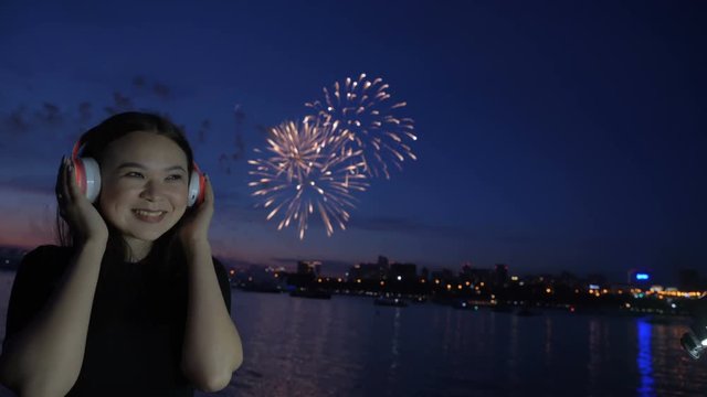 One young woman smile and listening music at headphones at blur scene with exploding fireworks at horizon slow motion. Free 20s female person enjoying alone at night on the holiday on the town beach