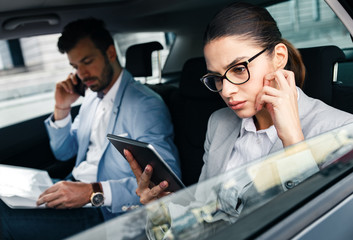 Young business people working together while traveling by a car. Businesswomen looking at tablet while businessmen talking at phone.