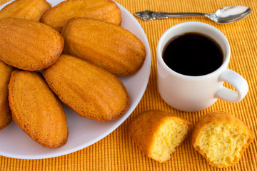 Delicious madeleine, homemade traditional french pastry (small cookie) on yellow tablecloth. Cup of coffee, spoon, halves of madeleine.