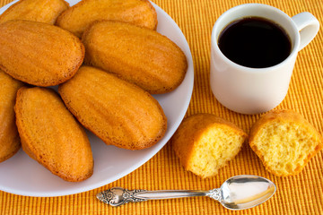 Delicious madeleine, homemade traditional french pastry (small cookie) on yellow tablecloth. Cup of coffee, spoon, halves of madeleine.