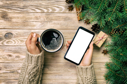 Female hands holding modern smartphone with mosk up and mug of coffee on wooden vintage table with christmas decoration. top view