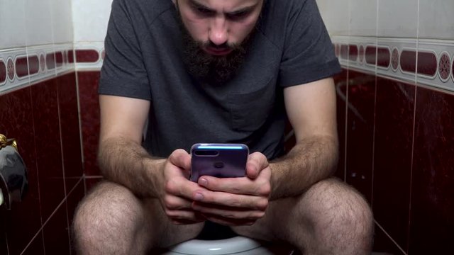closeup of a young caucasian man sitting in the toilet sending a message with his smartphone. Close up of a young caucasian man using his smartphone in the bathroom