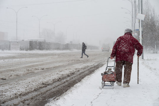 A snowstorm in the city. An elderly woman with a cane and a bag on wheels goes along the road during a snowstorm. Selective focus.