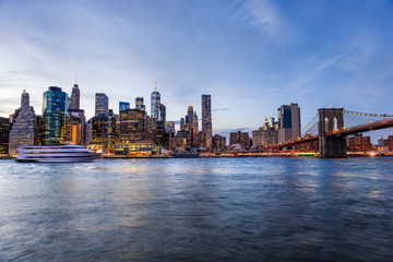 Fototapeta na wymiar Outdooors view on NYC New York City Brooklyn Bridge Park by east river, cityscape skyline at sunset, dusk, twilight, blue hour, dark night, skyscrapers, buildings, waves, tour boat