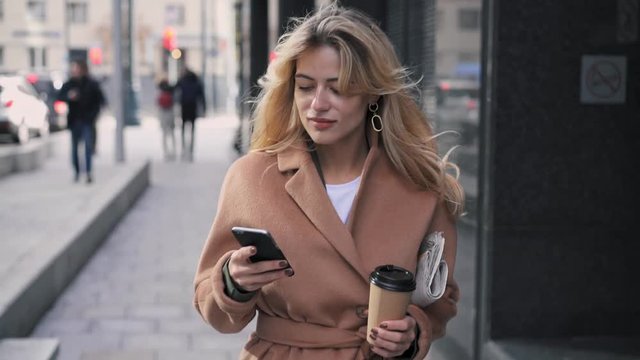 Attractive blonde woman wearing beige coat, holding a cup of coffee and a newspaper walking in city street and web surfing. Tracking slow motion medium shot