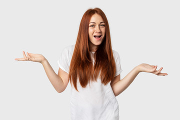 Young emotional European female isolated on grey background. Red-haired girl smiles in a white T-shirt. Shows surprise with hand gestures