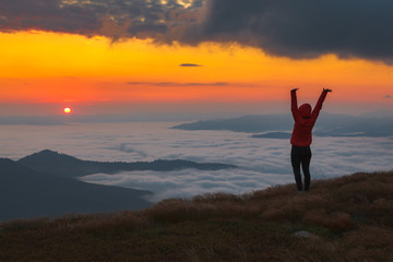 A girl in a red jacket with a hood watching from the top of the mountain sunrise over a mist-covered valley