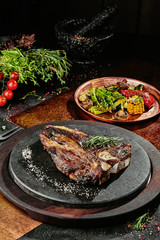 Fermented steak. T-Bone steak serving with grilled vegetables on a black round plate on a background of greens, spices and vegetables, on a copper sheet. close up