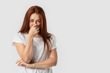 Young redhead European female isolated on white background dressed in white t-shirt, covering nose...