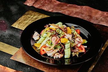 salad with ham, orange slices, tomato, cucumber and lettuce leaves in a black plate on a copper background. close up