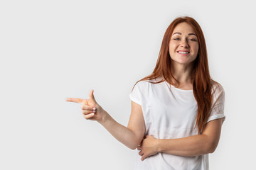 Beautiful young redhead female pointing left with finger isolated on gray background