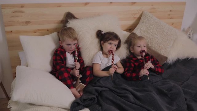 Three young children in red pajamas lie in a bed in a cozy living room and eat sweet candy on a stick. Christmas concept. Home holiday