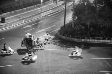 Man riding tricycle transporting a mountain of old staff and furniture. In the middle of a crossroads in China. Black and white photo.