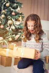 Happy Holidays. Cute little baby opens a Christmas present. Girl happy gifts sitting at the Christmas tree.