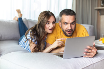 Fototapeta premium Surprised young guy and girl are watching something on gadget while lying on the couch