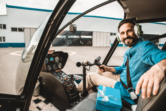 Positive emotional young man in colorful clothes sitting in the helicopter cabin and putting one hand on the seat while smiling and looking at you