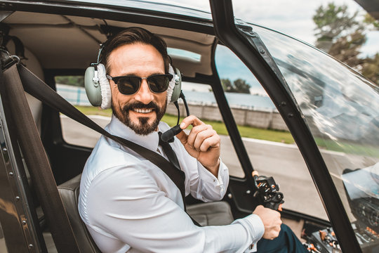 Positive enthusiastic young pilot sitting in the helicopter cabin and smiling while touching the headphones
