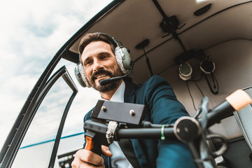 Attractive young man elegant pilot wearing big headphones and smiling while sitting in the helicopter cabin