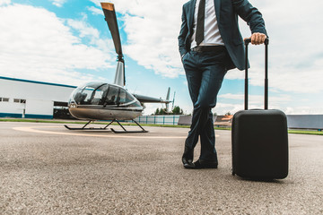 Elegant businessman standing on the helicopter platform and putting one hand on the rolling bag - Powered by Adobe