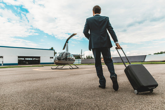 Business flight. Young businessman in dark suit walking with his luggage on the helicopter platform towards his helicopter
