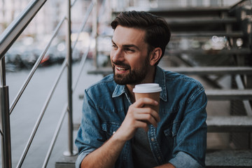 Portrait of happy male dreaming while tasting cup of delicious coffee. He sitting on stairs at street during rest