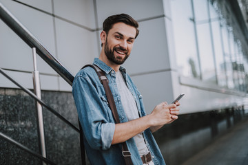 Portrait of outgoing bearded male typing in mobile while looking at camera outdoor