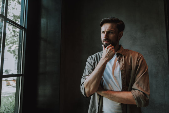 Portrait of pensive unshaven male touching chin with hand while looking at street in apartment