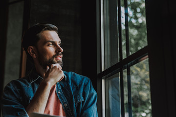 Side view thoughtful bearded man keeping chin with hand while looking at window during relax in room