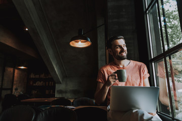 Fototapeta na wymiar Portrait of beaming bearded man tasting cup of coffee while looking at window indoor. He having job with digital device