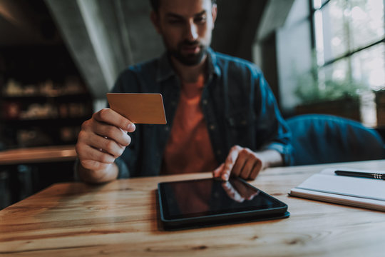Concentrated unshaven male typing in electronic tablet while demonstrating credit card in hand. He sitting at table indoor