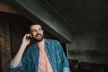Portrait of optimistic unshaven male talking on mobile while locating indoor. Glad guy using...