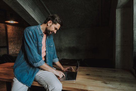 Side view glad bearded man using contemporary laptop while sitting on table in apartment. Happy dude during job with appliance concept