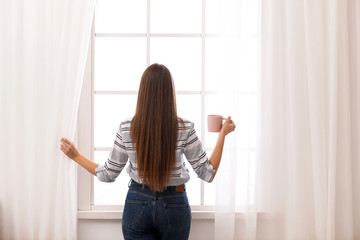 Young woman with cup of coffee opening window curtains at home