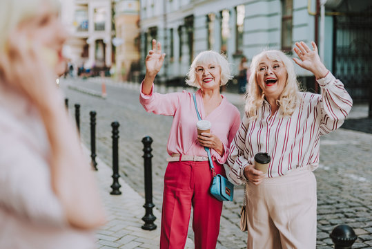 Lovely photo of smiling females in handsome clothes. Ladies standing on the street and waving to friend
