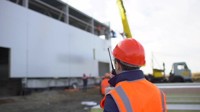 Male engineer, builder, foreman, worker in orange a uniform and hard hat using a walkie talkie giving instructions at a construction site. installation of sandwich panels of unfinished building