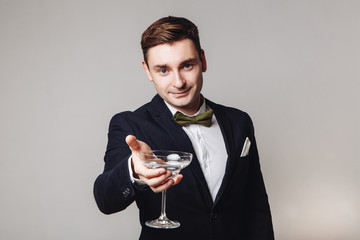 Young handsome man in black jacket and bow tie holding a drink and making toast gesture isolated on grey background. New year's eve. Offering a drink