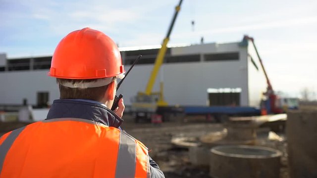 Male engineer, builder, foreman, worker in orange a uniform and hard hat using a walkie talkie giving instructions at a construction site. installation of sandwich panels of unfinished building